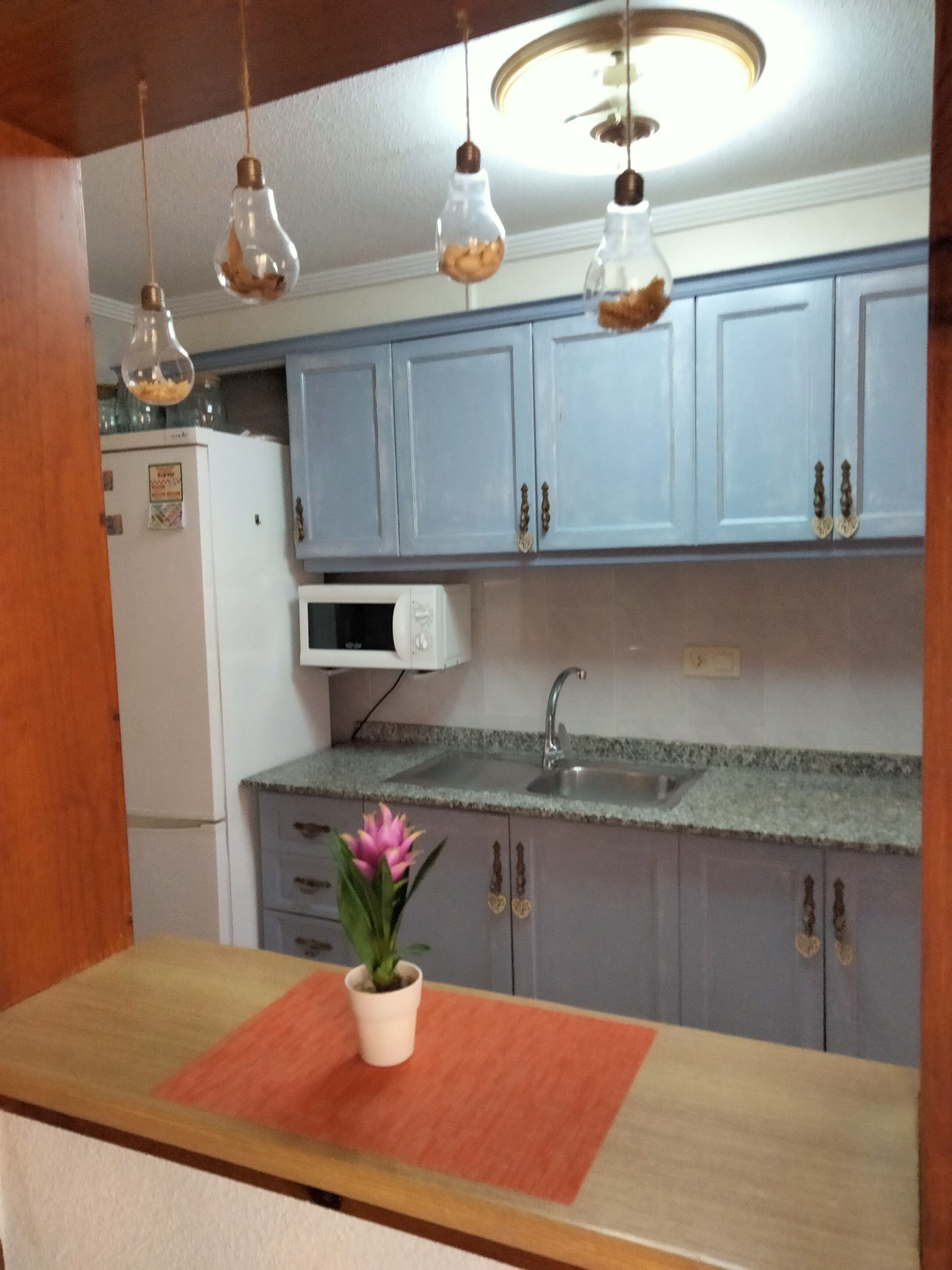 Une cuisine avec des meubles de couleur bleu clair, un réfrigérateur blanc, un four à micro-ondes, un évier et des luminaires suspendus avec des ampoules.