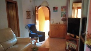 Ein Wohnzimmer mit einer Couch, einem Couchtisch mit einer Pflanze, einem blauen Sessel, einem Fernseher und einem Bücherregal.