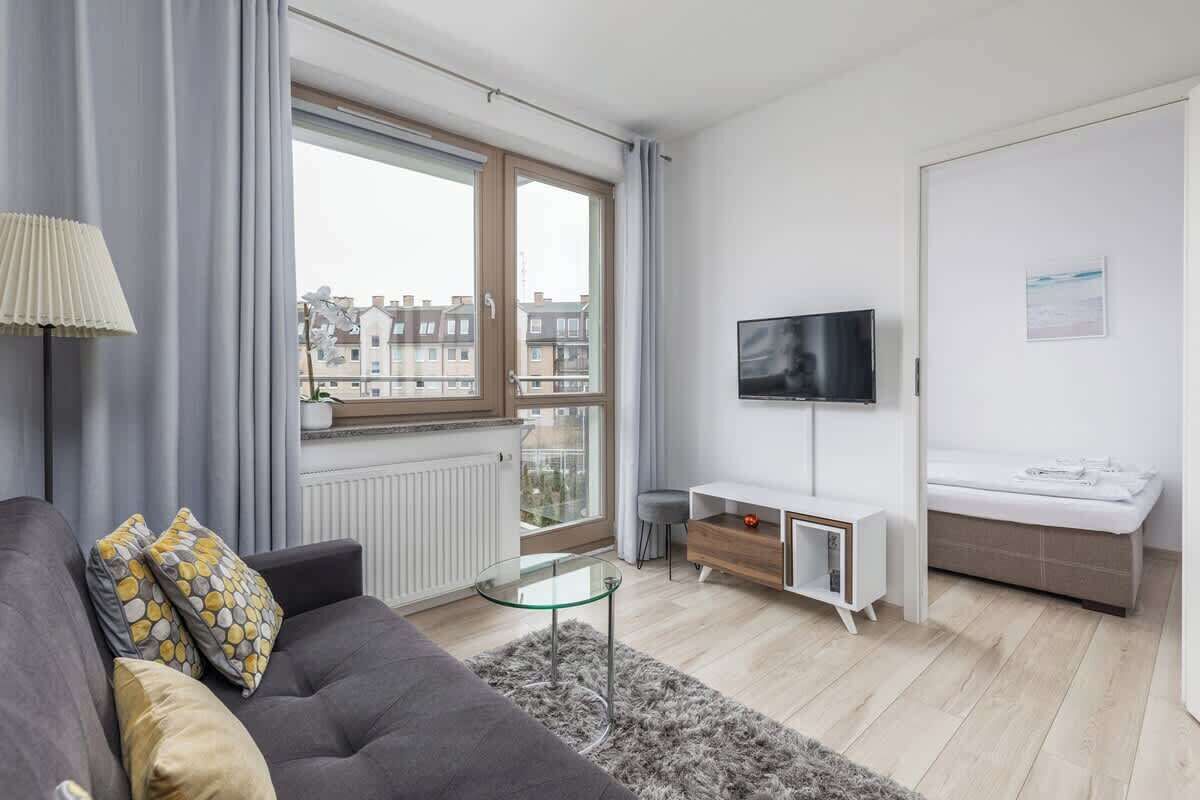 Ein modernes Wohnzimmer mit grauem Sofa, einem Glastisch, einem an der Wand befestigten Fernseher, einer kleinen weißen Kommode und einem Fenster mit Blick auf Gebäude.