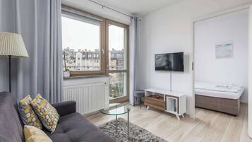 Ein modernes Wohnzimmer mit grauem Sofa, einem Glastisch, einem an der Wand befestigten Fernseher, einer kleinen weißen Kommode und einem Fenster mit Blick auf Gebäude.