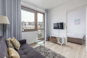 Ein modernes Wohnzimmer mit grauem Sofa, einem Glastisch, einem an der Wand befestigten Fernseher, einer kleinen weißen Kommode und einem Fenster mit Blick auf Gebäude.
