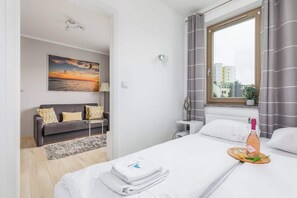 Hotellihuone, jossa on sänky, yöpöytä, verhoilla varustettu ikkuna, koristetyynyillä varustettu sohva, taulu seinällä ja samppanjapullo sängyllä.