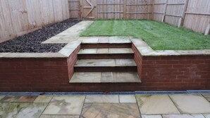 A backyard with a stone patio, a brick retaining wall, a wooden fence, and a grassy lawn. - 4 Bedroom Farnborough Airport Accommodation (Hampshire)