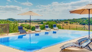 Ein klares, blaues Schwimmbecken mit Liegestühlen und Sonnenschirmen, umgeben von einem Zaun und Grünflächen, mit Blick auf eine entfernte Landschaft.