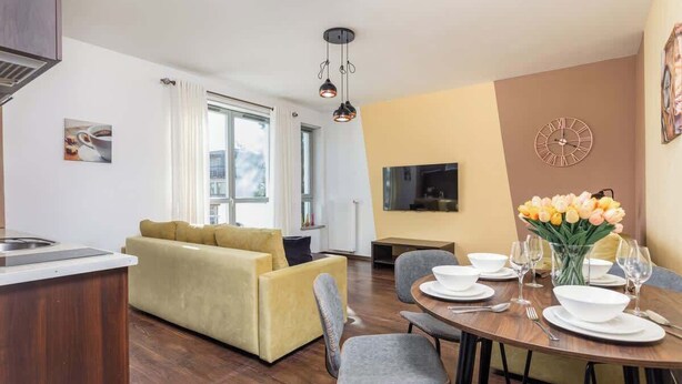Una sala de estar moderna con un sofá amarillo, comedor con una mesa redonda para cuatro personas, y una cocina con estufa.