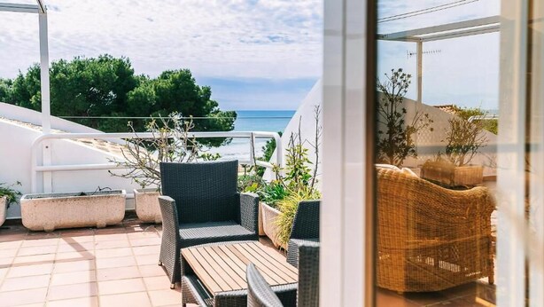Un balcon agrémenté de meubles en osier et de plantes en pot, offrant une vue sur une étendue d’eau et sur la verdure.