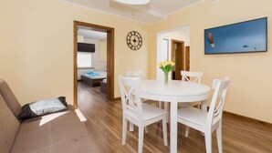 Ein modernes Wohnzimmer mit einem runden Esstisch, weißen Stühlen, einer Couch mit gemustertem Kissen, einer Wanduhr und einem flachbildfernseher an der Wand.