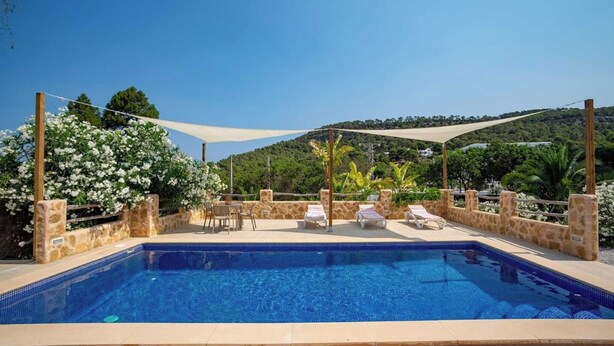 Una piscina rectangular con bordes azulejados, rodeada de muros de piedra y una pérgola de madera con una vela de sombra blanca.