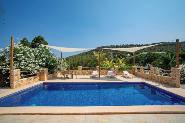 A rectangular swimming pool with tiled edges, surrounded by stone walls and a wooden pergola with a white shade sail.