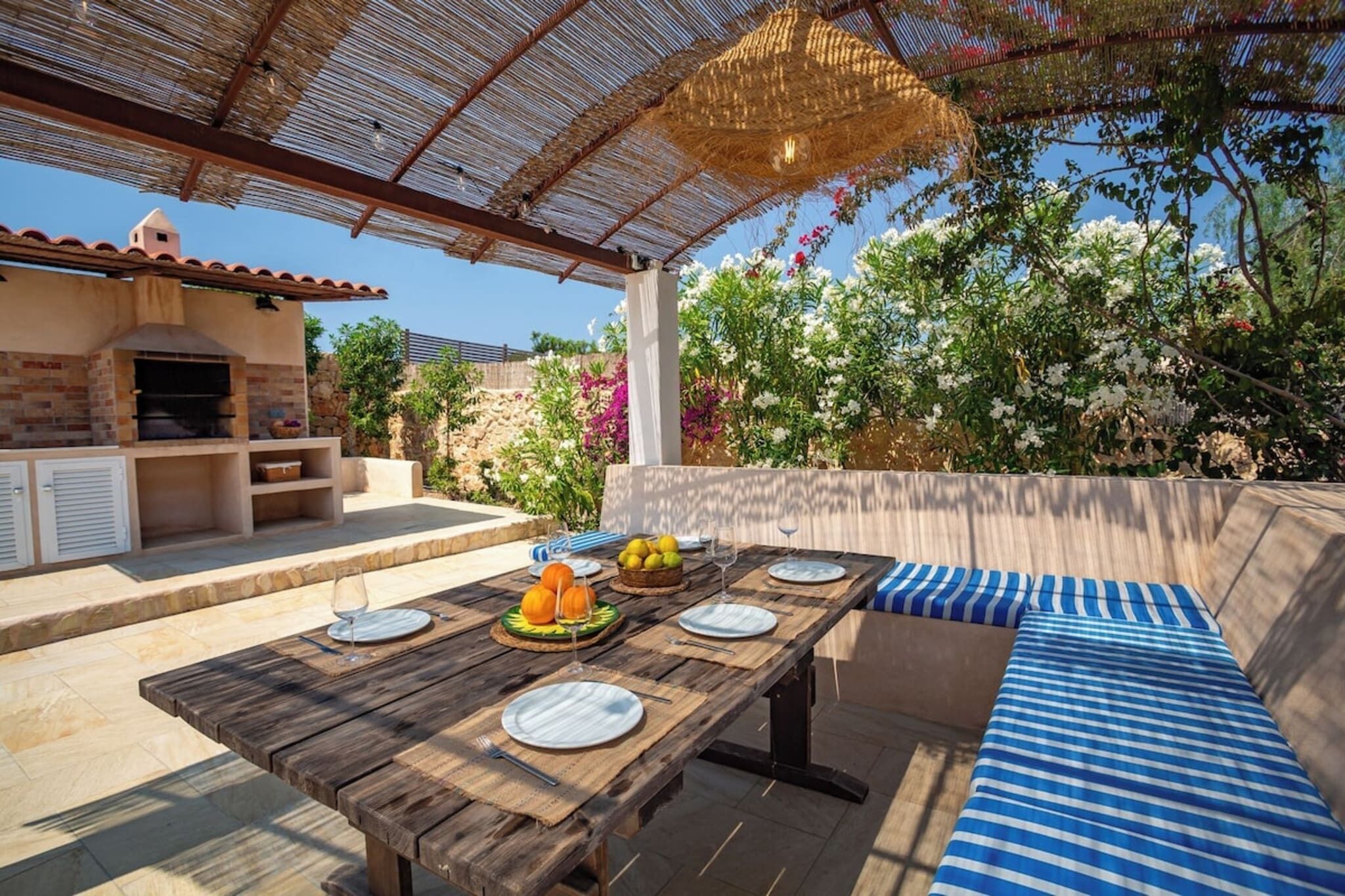Un espacio al aire libre con comedor, una mesa de madera dispuesta para cuatro, un horno de ladrillo y un muro de piedra de fondo.
