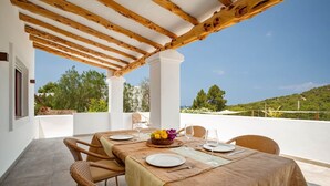 Un área de comedor al aire libre cubierta con una mesa lista para cuatro personas, rodeada de sillas de mimbre, bajo una pérgola de madera.