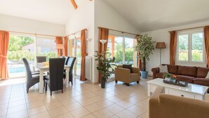 Ein geräumiger Wohnbereich mit Esstisch, Stühlen, einer Couch und einem Couchtisch. Große Fenster mit Vorhängen, ein Topfpflanze und ein Blick ins Freie.