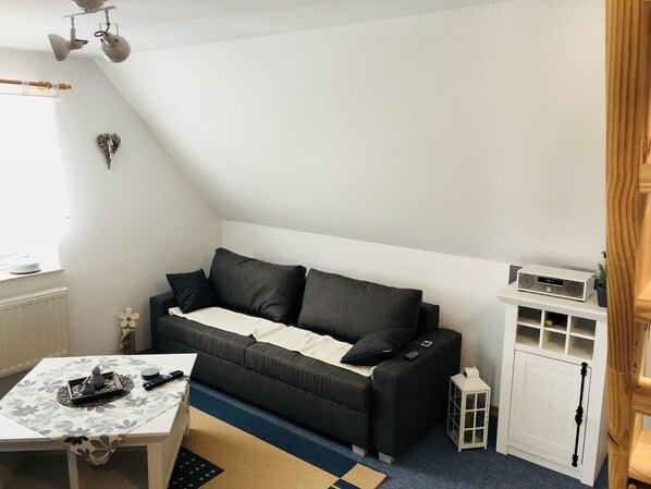 A small, well-lit room with a dark grey sofa, a white cabinet with a radio, a small table with a decorative tray, and a window with a curtain.