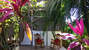 A wooden deck with two rocking chairs, surrounded by lush tropical plants and a large window. - Pequeñito Paraíso by LEELOU (Jacó)