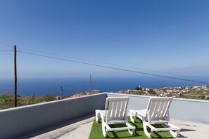 Two white lounge chairs on a rooftop terrace with a view of the sea and a distant town.