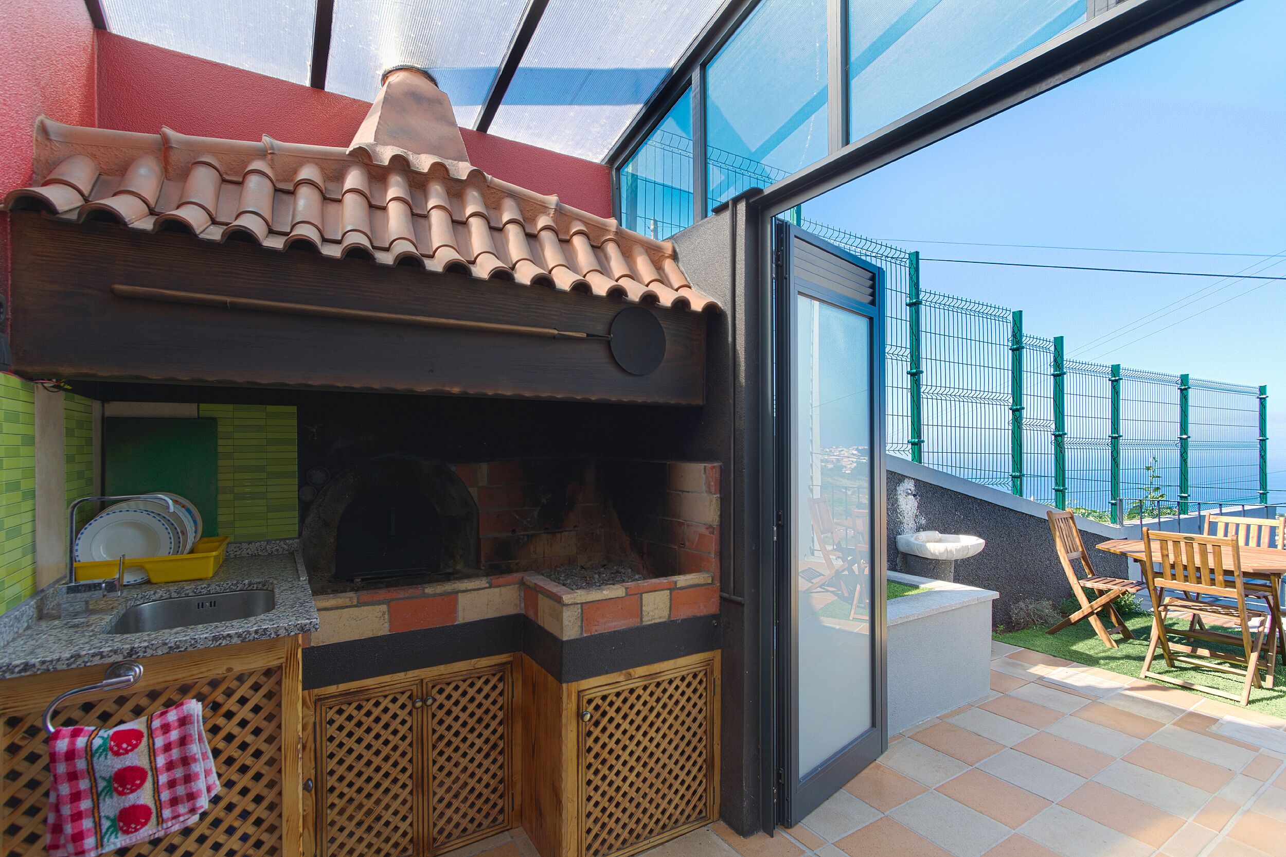 Una cocina al aire libre con horno de ladrillo, una mesa de comedor con sillas y vista al mar a través de un ventanal.