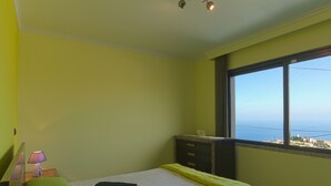 Une chambre à coucher avec un lit, des oreillers, une table de chevet, une lampe, une commode et une fenêtre donnant sur un beau paysage.