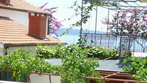 Una terraza con mesa y sillas, una planta en maceta con flores rojas, y vista al mar y edificios.