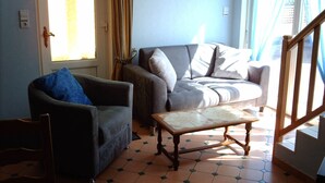 A living room with a sofa, armchair, and a small table. There is a window with curtains, a door, and a staircase. - Detached house "Chez Jean-Yves" with veranda and garage  (Le Menil)