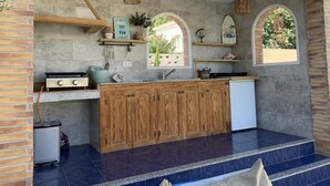 Een keuken met een centraal eiland, houten kastjes, een gasfornuis en een kleine koelkast. Er zijn twee ramen met gebogen bovenkanten, een hangende lamp en een plank met decoratieve objecten.