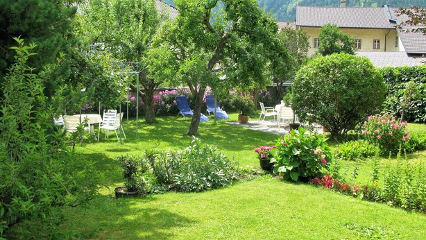 Un jardin luxuriant, avec une grande variété de plantes et de fleurs, un espace aménagé avec des chaises, et un bâtiment visible au fond.