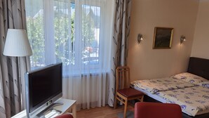Une chambre d’hôtel avec une télévision posée sur une table blanche, un lit recouvert d’une couvre-lit à motifs floraux, deux chaises rouges, une chaise en bois et un tableau accroché au mur.