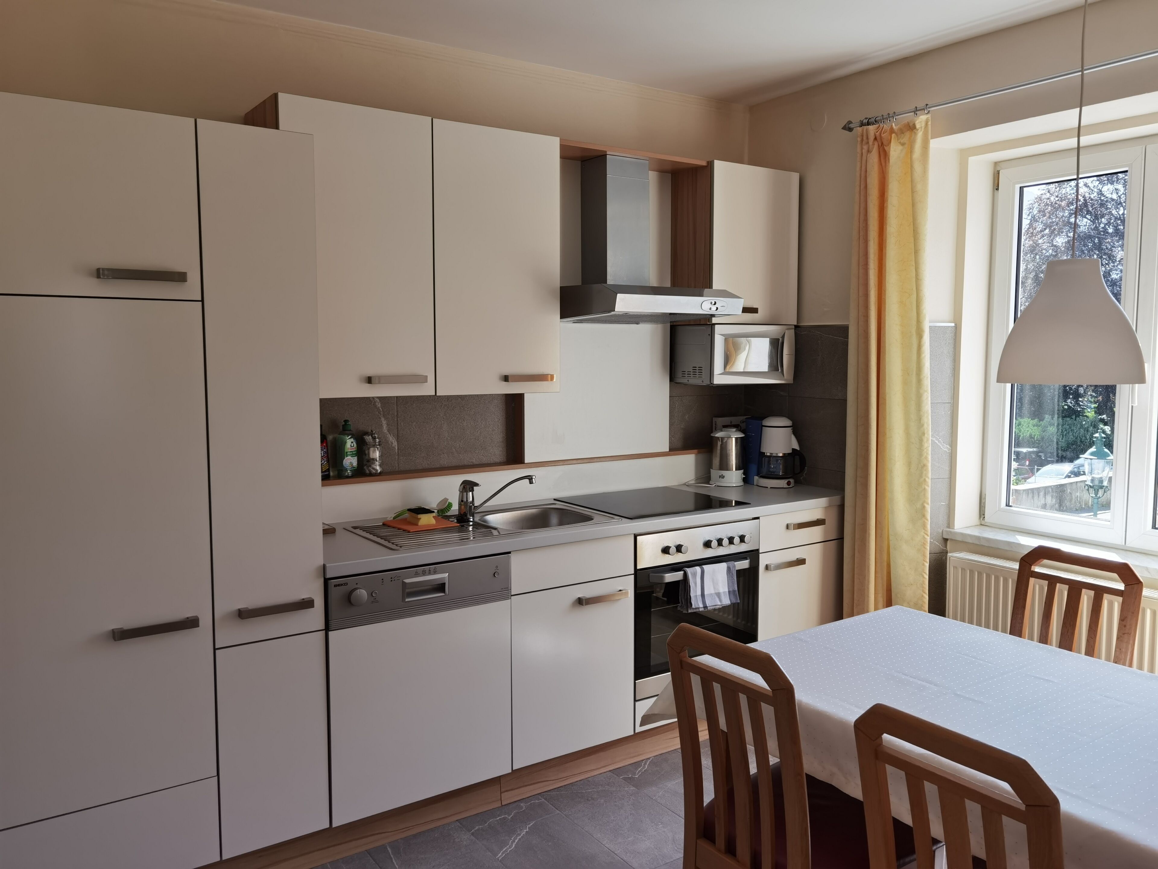 Una cucina moderna con mobili bianchi, forno a microonde integrato, piano cottura e lavello. C'è un tavolo da pranzo con sedie e una finestra con tende.