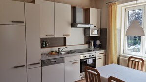 Une cuisine moderne avec des meubles blancs, un four à micro-ondes encastré, une cuisinière et un évier. On y trouve une table à manger avec des chaises et une fenêtre avec des rideaux.