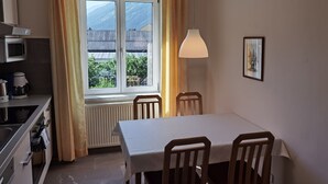 Une cuisine compacte équipée d’un four à micro-ondes, d’une machine à café et d’une cuisinière. Une table à manger avec quatre chaises est placée près d’une fenêtre ornée de rideaux.