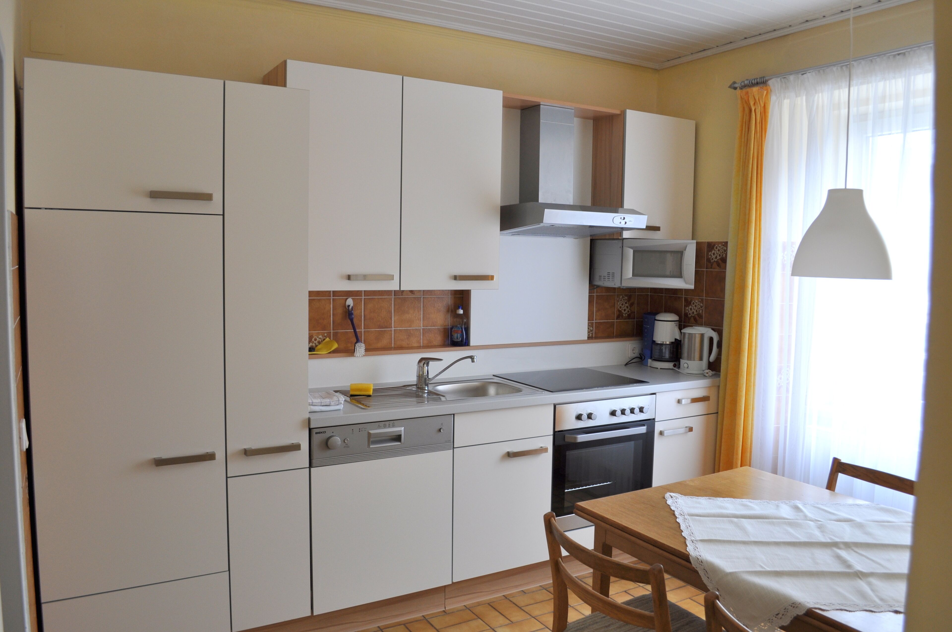 Eine kleine Küche mit weißen Schränken, einem Herd, einem Backofen und einer Mikrowelle. Ein Holztisch mit weißer Tischdecke und Stühlen steht daneben.