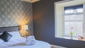 Una cama cuidadosamente hecha con sábanas blancas y toallas enrolladas, una lámpara de noche, un almohadón negro y una ventana con vistas a un edificio y una flor roja.