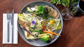 Un plato de verduras y hojas verdes sobre una mesa de madera, con un juego de tenedor y cuchillo sobre una servilleta a la izquierda.