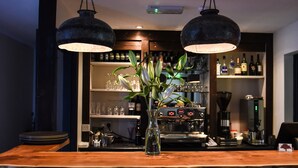 Una zona de bar con mostrador de madera, dos lámparas colgantes, un jarrón con flores, estantes con botellas y vasos, y una máquina de café.