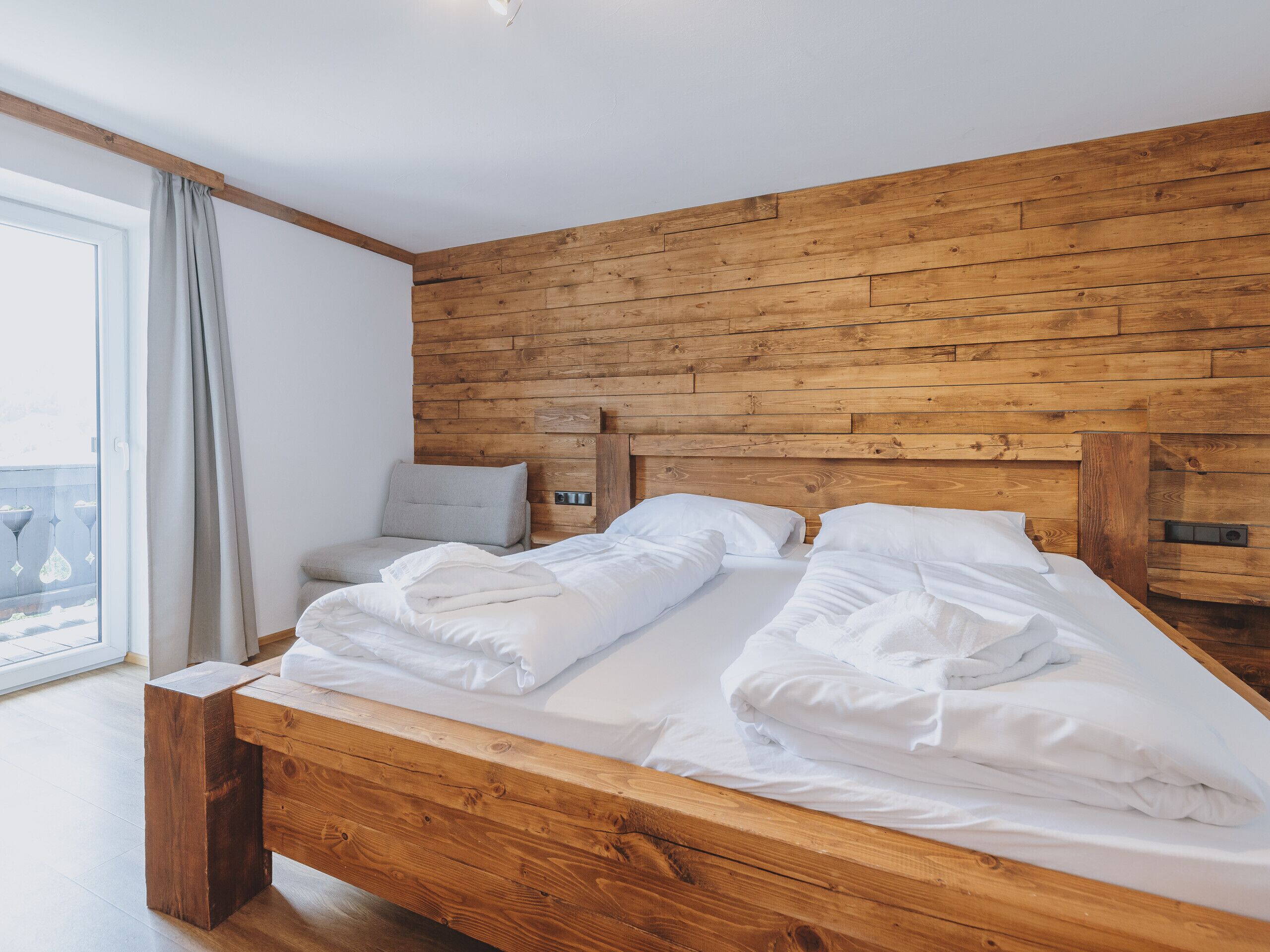 Een houten bed met witte lakens in een kamer met een houten hoofdbord en een groot raam met gordijnen.