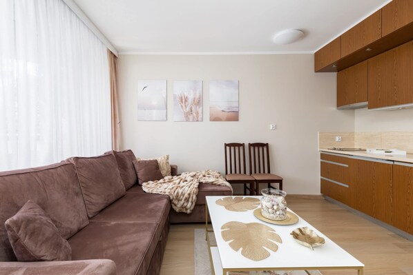 A modern living room with a brown sofa, a dining table with decorative items, and a kitchen area with wooden cabinets. - Cozy Apartment | Terrace, Parking, Close to the Beach (Świnoujście)