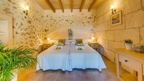 Un lit double avec une couvre-lit blanche, deux tables de chevet avec des lampes, une tête de lit en bois, un mur décoré d’un motif imitant la pierre et une plante en pot.