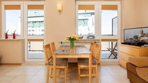 A dining area with a wooden table, chairs, and a vase of flowers. There are windows with blinds, a television, and a sofa. - Cozy Studio close to the Beach | Sauna and Gym (Kolobrzeg)