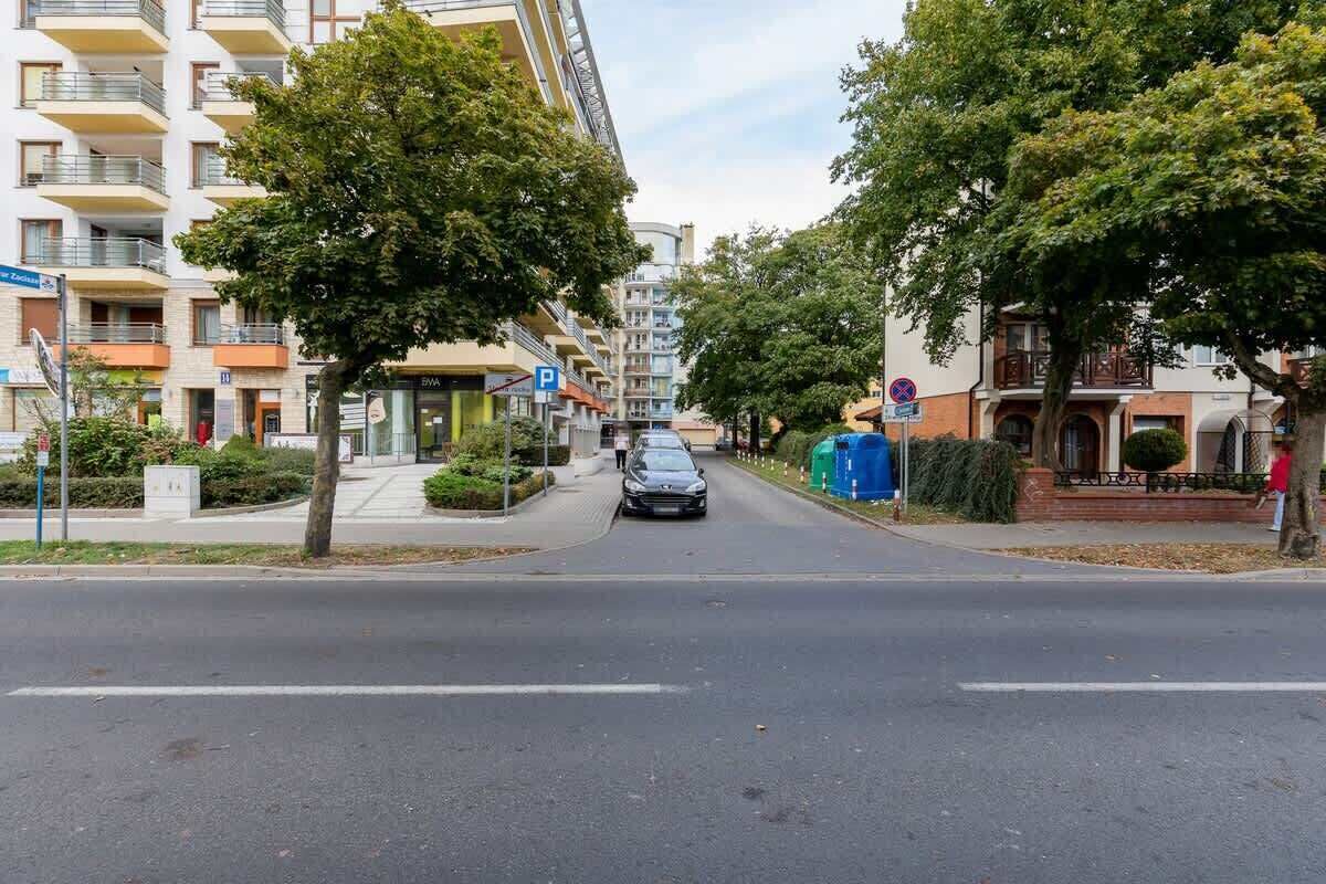A street with a car driving down, trees lining the road, and buildings with balconies and windows.