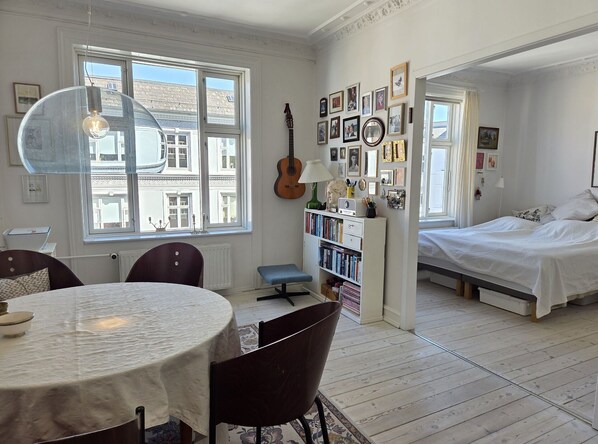 A room with a dining table, chairs, a bookshelf, a guitar, and a bed.