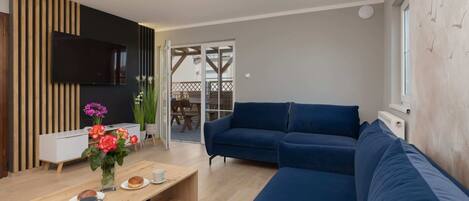 Una sala moderna con un sofá modular, un televisor de pantalla plana montado en un panel de madera, una mesa de centro con un jarrón de flores y un portón corredizo de vidrio que da a un espacio exterior.