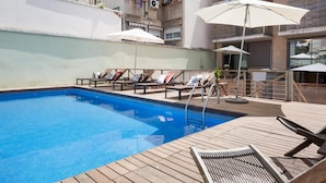 A rooftop pool with wooden loungers and umbrellas, surrounded by a high-rise building with balconies.
