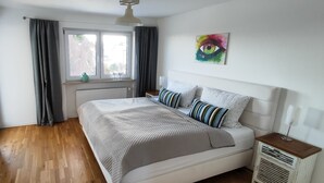A bedroom with a bed, a window, a radiator, and a wall with a painting. - Cozy house with garden / near Simssee (Stephanskirchen)