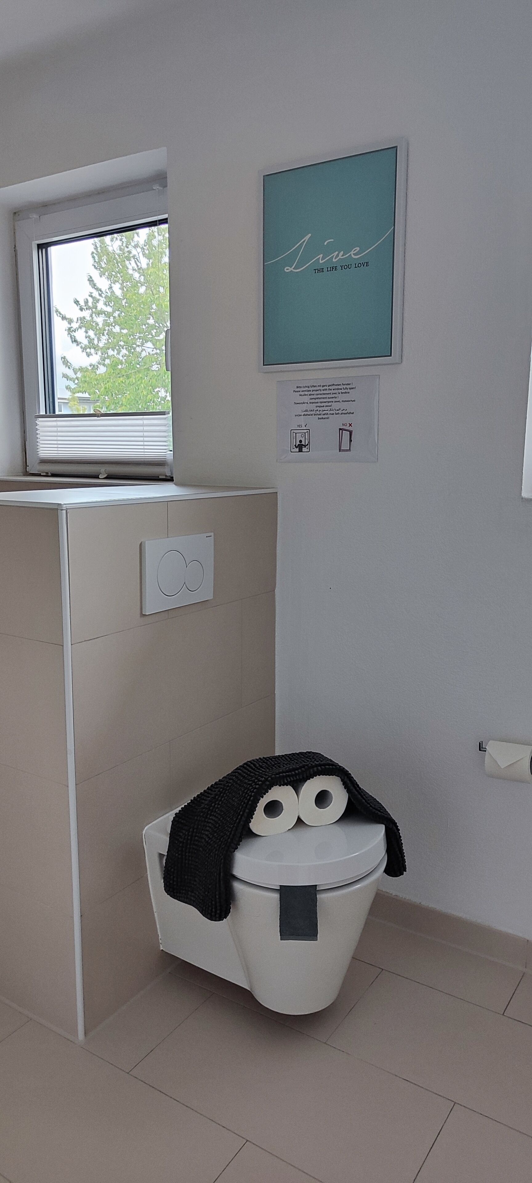 Ein Badezimmer mit Toilette, einem Fenster, einem Thermostat und einem gerahmten Motivationszitat an der Wand.