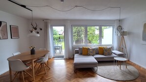 A modern living room with a wooden herringbone floor, a grey sofa with patterned cushions, a round coffee table, and a dining area with a wooden table and chairs. - Cozy house with garden / near Simssee (Stephanskirchen)