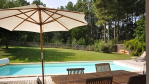 Eine Holzterrasse mit Billardtisch, Stühlen, einem großen Sonnenschirm und einem Schwimmbad.