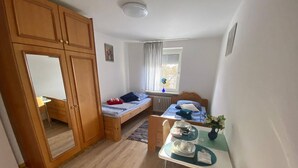 Ein gemütlicher Raum mit einem Bett, einem Schreibtisch und einem Sessel. Ein Fenster mit Vorhängen, ein Heizkörper und ein kleiner Tisch mit einer Vase.