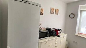Eine kompakte Küche mit einem weißen Kühlschrank, einer Mikrowelle, einer Kaffeemaschine und einem Toaster auf einem weißen Schrank. An der Wand hängen drei Bilder und eine Uhr.