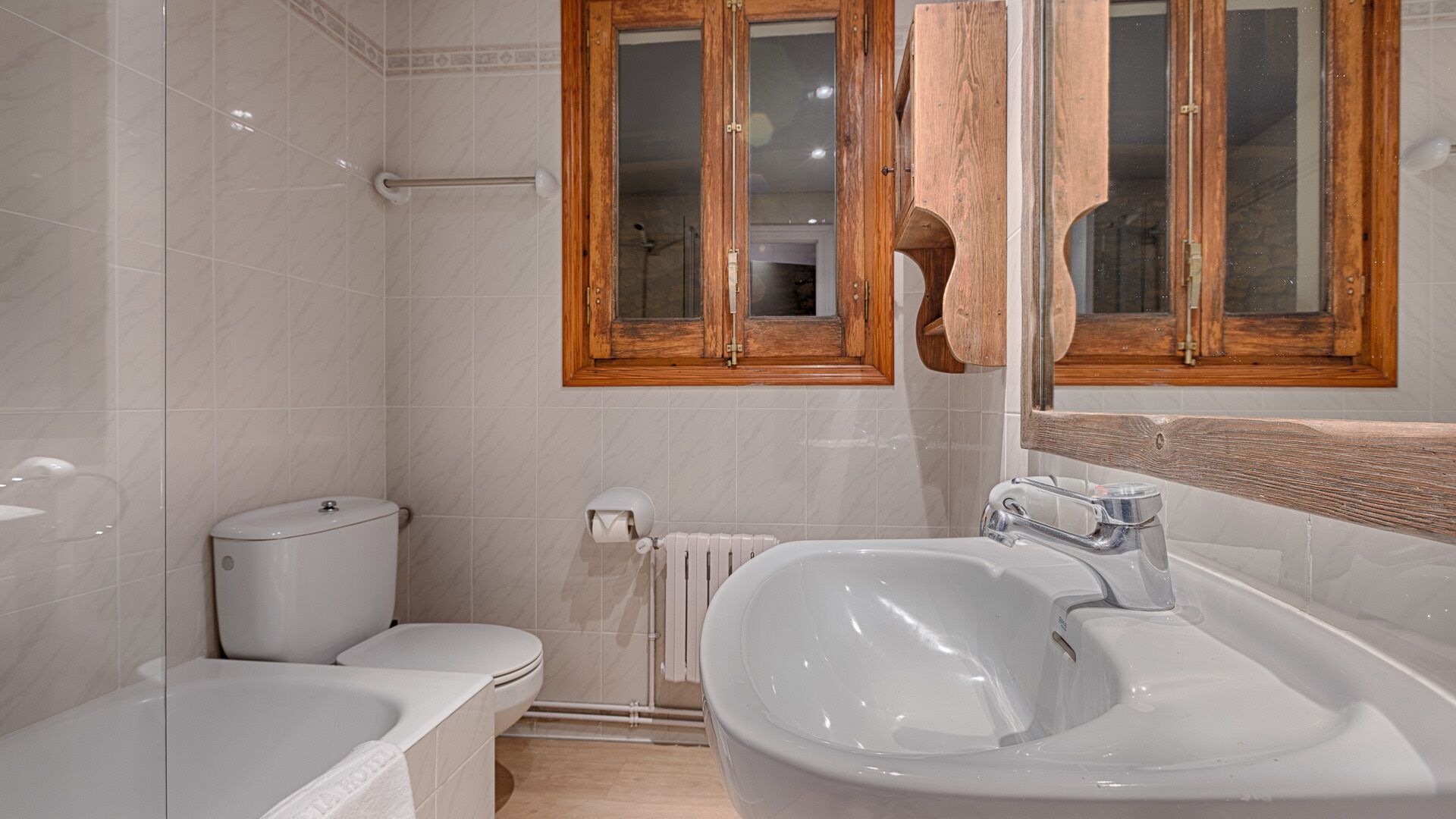 Un baño moderno con bañera, lavamanos e inodoro. Hay armarios de madera sobre el lavamanos y una ventana sobre el inodoro.