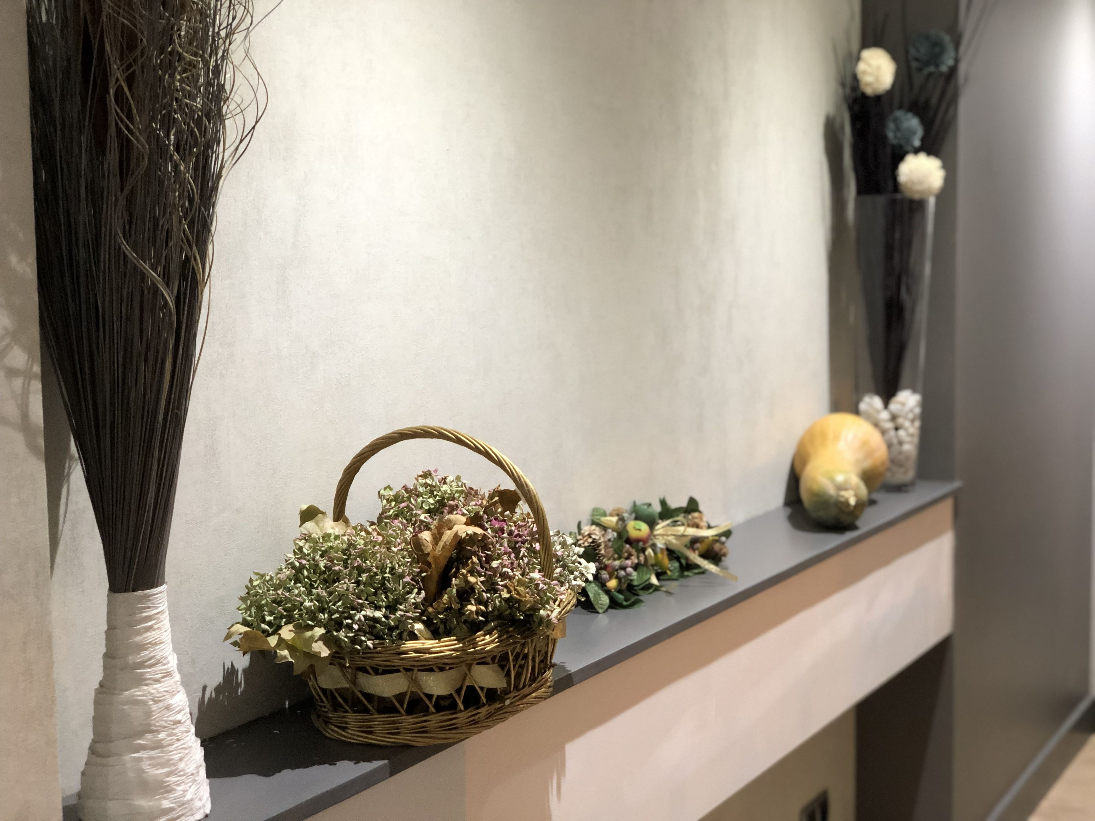 A shelf with a wicker basket containing dried flowers and foliage, a white vase with a tall, dark plant, and a decorative ceramic piece.