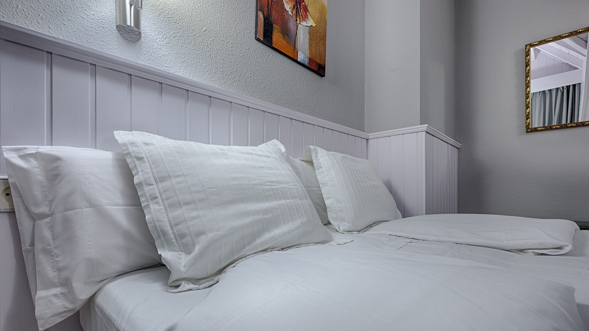 A neatly made bed with white linens and pillows in a room with white paneled walls and a painting above the bed.